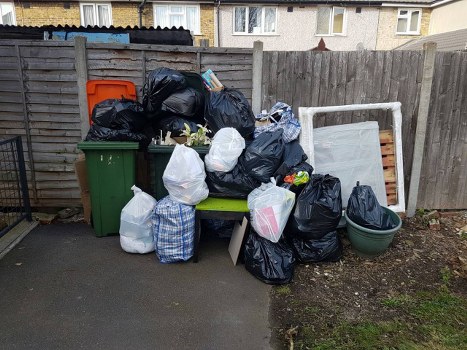 A collection of black, white, and blue rubbish bags filled with waste materials is piled on a paved driveway, with some bags placed on a small green bench situated against a wooden fence. Additional waste includes a large green wheelie bin, a smaller black bin, and a green plastic container, all positioned nearby. A dismantled metal bed frame with a white finish leans against the fence on the right side of the image, with a section of garden soil visible at the edge of the paved area. The background features residential buildings with multiple windows and a grey exterior finish, suggesting an urban or suburban environment. The scene appears to be part of a garden clearance service, with the rubbish bags ready for removal, possibly by Garden Clearance Woolwich. The lighting is natural, indicating daytime, and the setting is an outdoor space within Woolwich, supporting local rubbish removal activities.