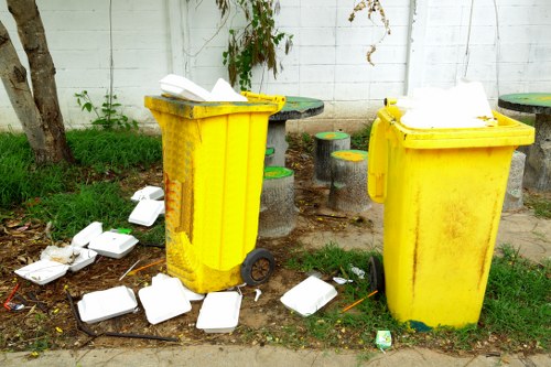 Two yellow wheeled rubbish bins made of plastic with textured surfaces are positioned on a grassy and paved area outdoors next to a white painted wall. The bin lids are partially open, revealing white foam containers inside. Several foam containers and pieces of debris are scattered on the ground around the bins, some leaning against the bins and others lying flat. Small concrete stools with green and yellow tops are arranged nearby, situated behind the bins, with parts of a garden fence or similar structure partly visible in the background. The scene is illuminated by natural daylight, and the environment appears to be a garden or outdoor space designated for waste disposal, consistent with rubbish removal services in Woolwich.