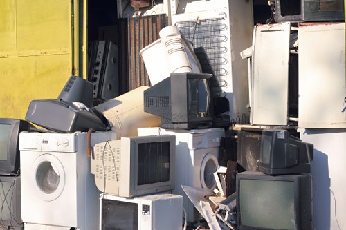 A large pile of discarded electronic waste and household appliances stacked outdoors on a paved surface, including washing machines, old computer monitors, and various electronic units with visible screens and control panels. The appliances are arranged irregularly, some leaning against each other, with different sizes and finishes, mostly in shades of white, black, and grey. In the background, there are industrial storage units or containers, partly obscured, with some metal surfaces and a glimpse of a yellow wall. The image captures a waste collection site or a rubbish removal operation, with natural daylight illuminating the scene, suggesting a typical outdoor yard or depot associated with waste clearance services like those offered by Garden Clearance Woolwich, serving the Woolwich area and surrounding postcode districts.