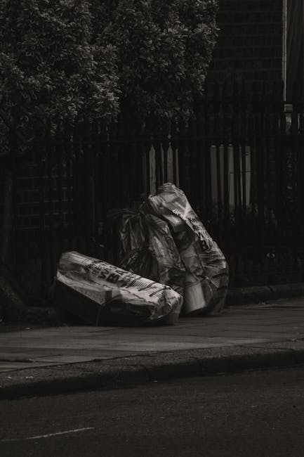 A black wheeled rubbish bin marked with 'St. John's' on the front, placed on a sidewalk next to a curb in an urban residential area at night. The bin is overflowing with mixed waste, including crumple